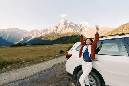 Счастливая женщина, опирающаяся на автомобиль на обочине дороги с горами и широким пейзажем, образ жизни путешественника и свобода путешествия на автомобиле, отдых на природе и улыбающаяся туристка…