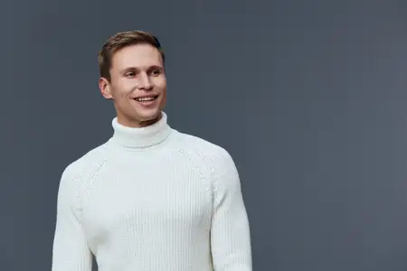 Счастливый молодой человек со светлой кожей и каштановыми волосами в уютном белом свитере стоит на нейтральном сером фоне, улыбается и смотрит в сторону, излучая тепло и дружелюбие
