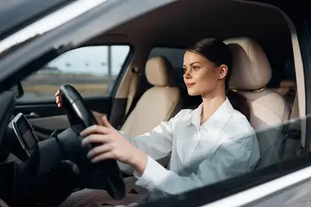 Уверенная женщина сидит на водительском сиденье автомобиля, держа руки на руле и глядя прямо в камеру со спокойным выражением лица