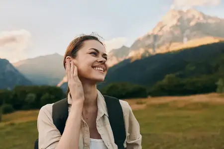 Молодая женщина с длинными каштановыми волосами наслаждается солнечным горным походом в зеленой долине, одетая в бежевую рубашку и белый топ, с черным рюкзаком за плечами, чувствуя себя расслабленной