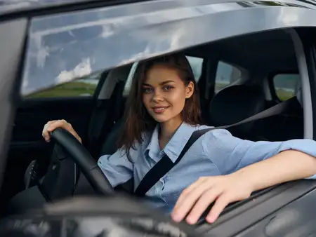 Молодая женщина на водительском сиденье автомобиля, рука на руле, смотрит прямо в камеру с нейтральным выражением лица