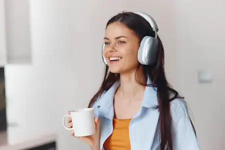 Женщина наслаждается музыкой в наушниках и кофе на кухне, одновременно пользуясь смартфоном