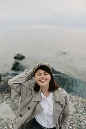 Счастливая женщина в уютной одежде, шляпе и пальто стоит на каменистом берегу у спокойного океана
