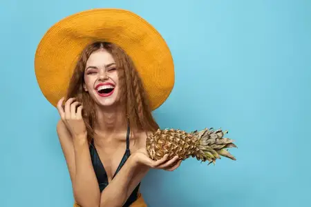 Женщина в пляжной шляпе держит ананас и купальник на фоне экзотических фруктов и голубого фона, передавая атмосферу отпуска
