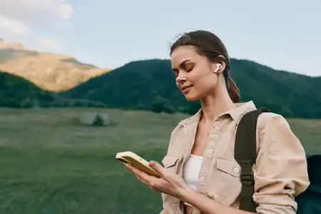 Молодая женщина в бежевой куртке и белом топе стоит в зеленом горном ландшафте, используя беспроводные наушники и глядя на смартфон с легкой улыбкой