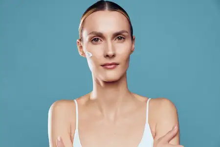 На ней белый топ при холодном освещении, синий фон подчеркивает чистоту и заботу о себе, пока она выполняет простой ритуал ухода за кожей