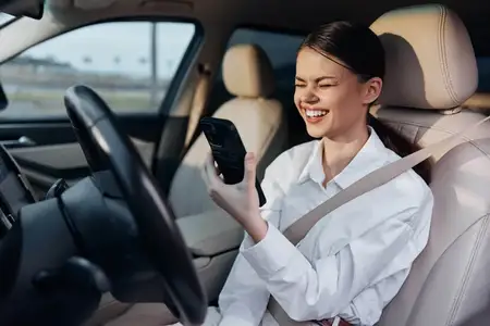 Женщина сидит за рулем автомобиля, прижимая телефон к уху и одновременно глядя на экран