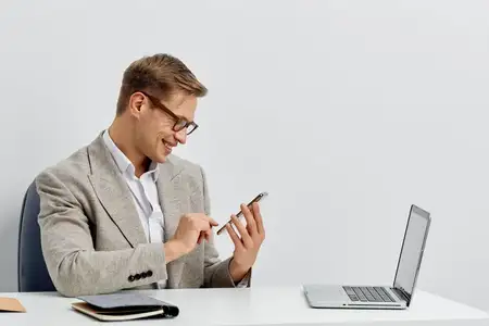 Улыбающийся мужчина работает со смартфоном и ноутбуком, в очках и повседневной куртке, сидит за столом на однотонном белом фоне, сосредоточен на кибербезопасности и концепции образа жизни цифрового…