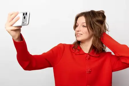 Улыбающаяся молодая женщина в красном свитере держит смартфон, чтобы сделать селфи, и естественно позирует, запустив руку в волосы, на белом фоне