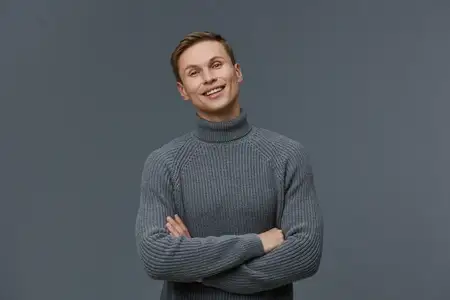 Счастливый молодой человек в уютном сером вязаном свитере с высоким воротником позирует, скрестив руки, и тепло улыбается в камеру