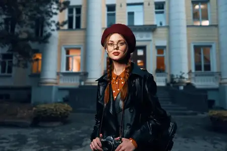 Уверенная женщина в красном берете и очках позирует на фоне современного городского здания, излучая стиль и утонченность
