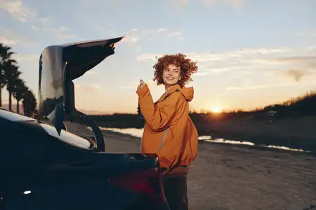 Образ жизни в поездке, на открытом воздухе, женщина стоит у открытого багажника машины на закате на пляже, вьющиеся волосы, повседневная куртка, багаж сзади, золотой свет, океанские волны