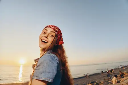 Улыбающаяся женщина с красной банданой стоит на залитом солнцем пляже во время заката, одетая в повседневный деним, с радостным и расслабленным настроением, на заднем плане море и люди, летняя…