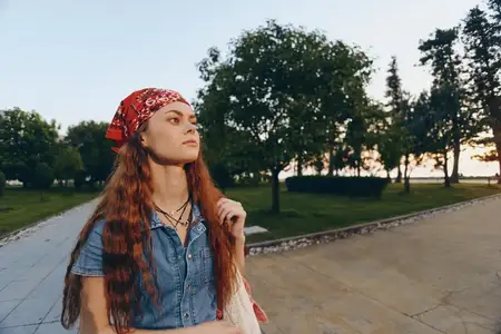 Женщина с длинными рыжими волосами в современном богемно-вестерн дениме и красной бандане идет на улице возле деревьев в теплом свете золотого часа, снятая в естественных пленочных цветах