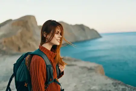 Симпатичная женщина с рюкзаком за спиной в горах у моря, вид сбоку, модель
