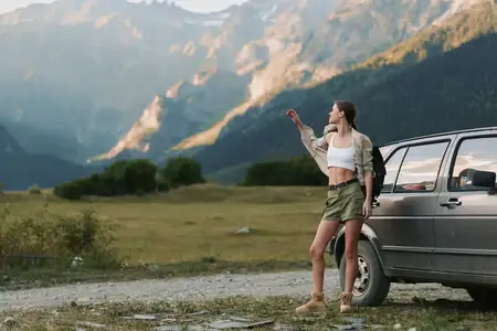 Молодая женщина в белом кроп-топе, зелёной юбке и лёгкой куртке стоит рядом с машиной на гравийной дороге в окружении гор и зелени, воплощая беззаботный момент жизни