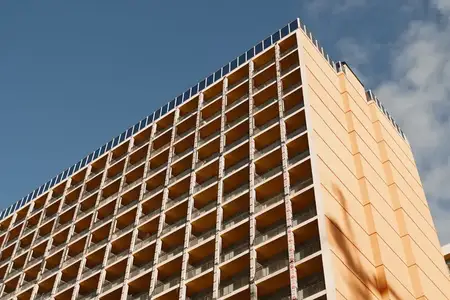 Архитектурная сцена: строящийся высотный дом с оранжевыми панелями и открытым каркасом, повторяющиеся решетчатые проемы, острые углы, яркое небо, акцент на современном дизайне
