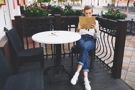 Стильная молодая женщина в рубашке сидит за столиком в летнем кафе с книгой в руках