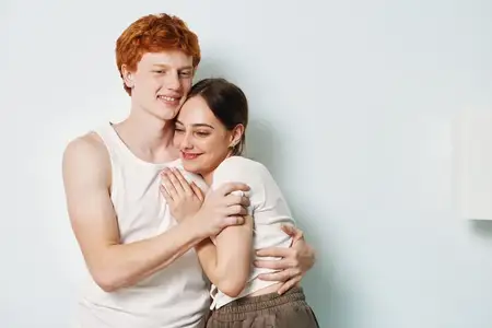 Счастливые молодые мужчина и женщина, тесно обнимающиеся с улыбками, в повседневной одежде, ласковая поза, эмоциональная связь и нежное прикосновение в светлой комнатной обстановке