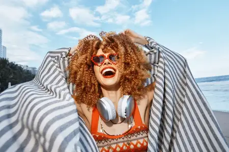 Солнечный пляжный отдых: улыбающаяся женщина наслаждается свободой и счастьем, у неё вьющиеся волосы и веснушки, она выражает эмоции, на ней стильные солнцезащитные очки и модные наушники, крупный…