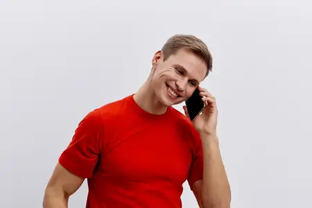 Счастливый мужчина в красной футболке держит мобильный телефон и улыбается, стоя на фоне однотонного белого задника