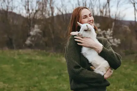 Естественная сцена на открытом воздухе показывает улыбающуюся женщину, обнимающую козлёнка на зелёных полях, подчёркивая деревенский образ жизни, товарищество, тепло и заботу между людьми и животными