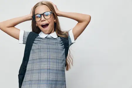 Счастливая школьница в форме и очках, держащаяся за лямки рюкзака, с радостным выражением лица и открытым ртом на однотонном белом фоне