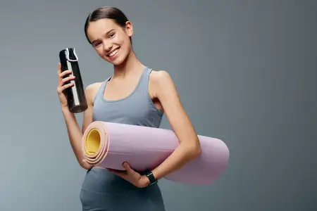 Спортивная женщина с ковриком для йоги и бутылкой воды готовится к упражнениям, подчеркивая фитнес-рутину, гибкость и осознанную тренировку в тренажерном зале или домашней студии