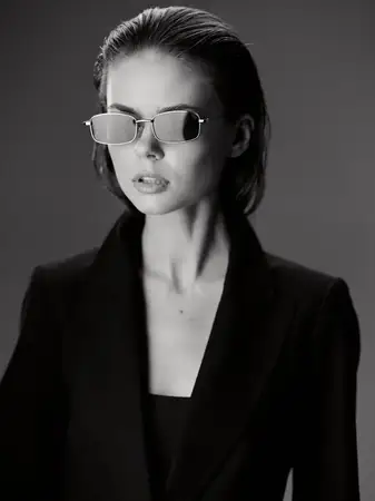 Черно-белый портрет модной женщины в солнцезащитных очках и черном блейзере с серьезным выражением лица на темном фоне, подчеркивающий элегантность и современный стиль