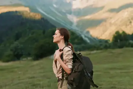 Молодая женщина, одетая в повседневную походную одежду, несёт большой рюкзак, стоя на зелёном лугу в окружении гор и наслаждаясь природным пейзажем и свежим воздухом