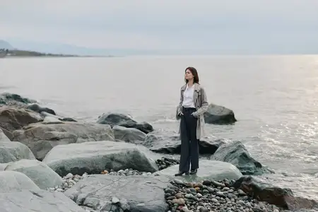 Задумчивая женщина в тренчкоте стоит на каменистом пляже у спокойной воды