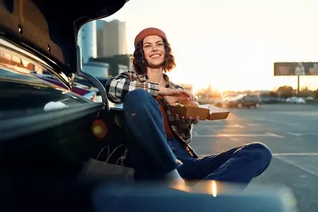 Непринужденная женщина рядом с автомобилем на парковке на закате держит еду и улыбается в городской сцене золотого часа, повседневный образ жизни с аутентичностью, осознанной жизнью и атмосферой…