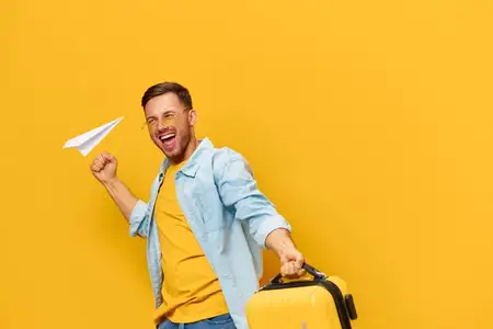 Невероятно радостный, веселый, загорелый, красивый мужчина в синей рубашке нетерпеливо ждет отпуска, держа чемодан, позируя изолированно на оранжево-желтом студийном фоне