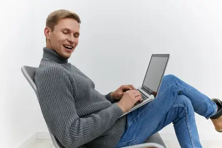 Улыбающийся мужчина работает за ноутбуком, одетый в уютный серый свитер и синие джинсы, сидит расслабленно на современном стуле на белом фоне, сосредоточен на образе жизни цифрового кочевника и…