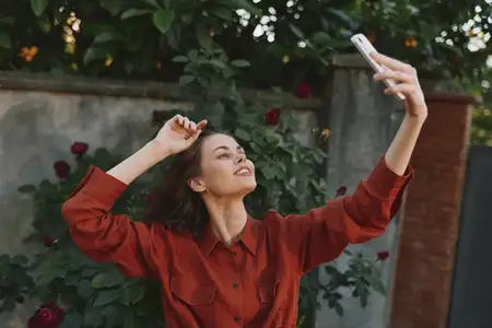 На сцене одна женщина в повседневной одежде позирует с телефоном среди зелени и цветущих кустов, ее теплая улыбка и расслабленная поза отражают естественный дневной свет и уверенность