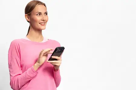Улыбающаяся женщина в розовом свитере держит смартфон, глядя в сторону на белом фоне