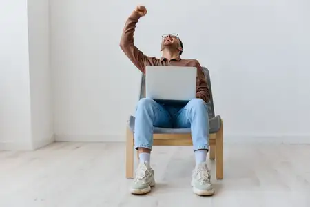 Удачливый, весёлый, невероятно радостный молодой загорелый мужчина с ноутбуком на коленях поднимает сжатые кулаки вверх, празднуя большую победу, и говорит «Да», сидя на стуле у белой стены дома