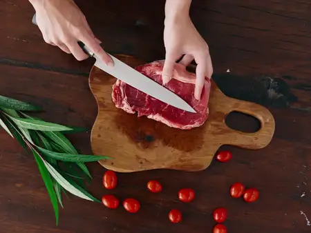 Женщина с ножом в руке нарезает свежий стейк для жарки на кухне, рядом на столе соль, перец и другие специи, красные помидоры черри и зелень