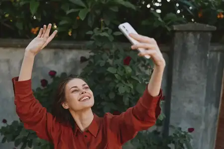 Улыбающаяся молодая женщина держит телефон над головой, чтобы сделать селфи на фоне пышного сада, излучая непринужденную уверенность и яркое, беззаботное настроение