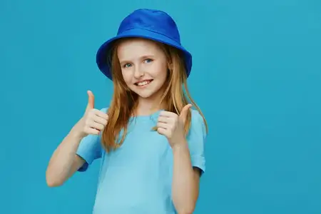 Счастливая маленькая девочка в синей шляпе и рубашке показывает большой палец вверх на фоне ярко-синего фона, олицетворяя радость и позитив, что идеально подходит для детских тем, мотивации и счастья