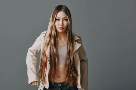 Привлекательная девушка с длинными прямыми волосами в повседневной одежде позирует на фоне серой стены