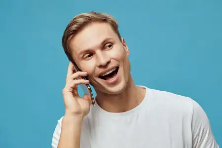 Портрет счастливого улыбающегося мужчины, разговаривающего по телефону, в белой футболке на голубом фоне