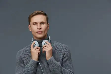 Серьезный молодой мужчина европеоидной расы в сером вязаном свитере с наушниками на шее смотрит в камеру со спокойным выражением лица, изолированный на однотонном сером фоне