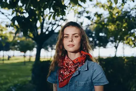 Молодая женщина в джинсовой рубашке и красной бандане стоит на улице в залитом солнцем парке среди деревьев и зеленой травы