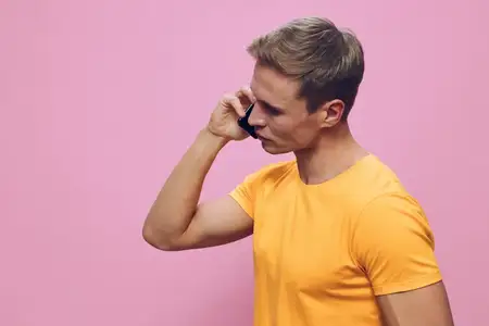 Молодой человек разговаривает по смартфону, одетый в желтую футболку, изолированный на розовом фоне, крупный план, вид сбоку, коммуникационные технологии, образ жизни цифрового кочевника, концепция…