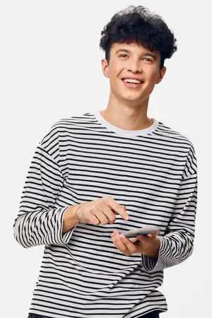 Молодой человек улыбается и пользуется смартфоном в повседневной полосатой рубашке на фоне однотонного белого фона, выражая радость и вовлеченность в мобильные технологии, концепция образа жизни