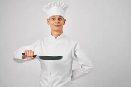 Мужчина в поварской одежде держит в руках сковороду и профессионально работает на кухне