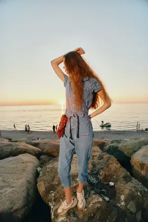 Женщина в стиле lifestyle, одетая в современный бохо-вестерн деним стритстайл, уверенно стоит на камнях у моря в золотой час, с мягким плёночным цветом и спокойным океанским фоном
