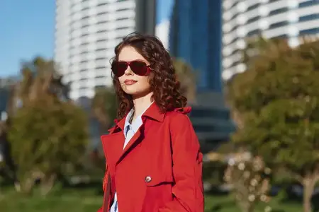 Привлекательная молодая женщина в красном пальто и тёмных солнцезащитных очках позирует на улице в городском парке с современными высотными зданиями и зелёными деревьями в яркий солнечный день