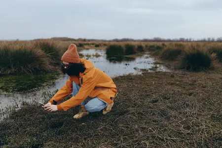 Женщина на открытом воздухе сидит и смотрит на болотную воду, в оранжевой куртке и капюшоне, трава на берегу реки и у пруда, облачное весеннее небо, безмятежный момент досуга на природе для…